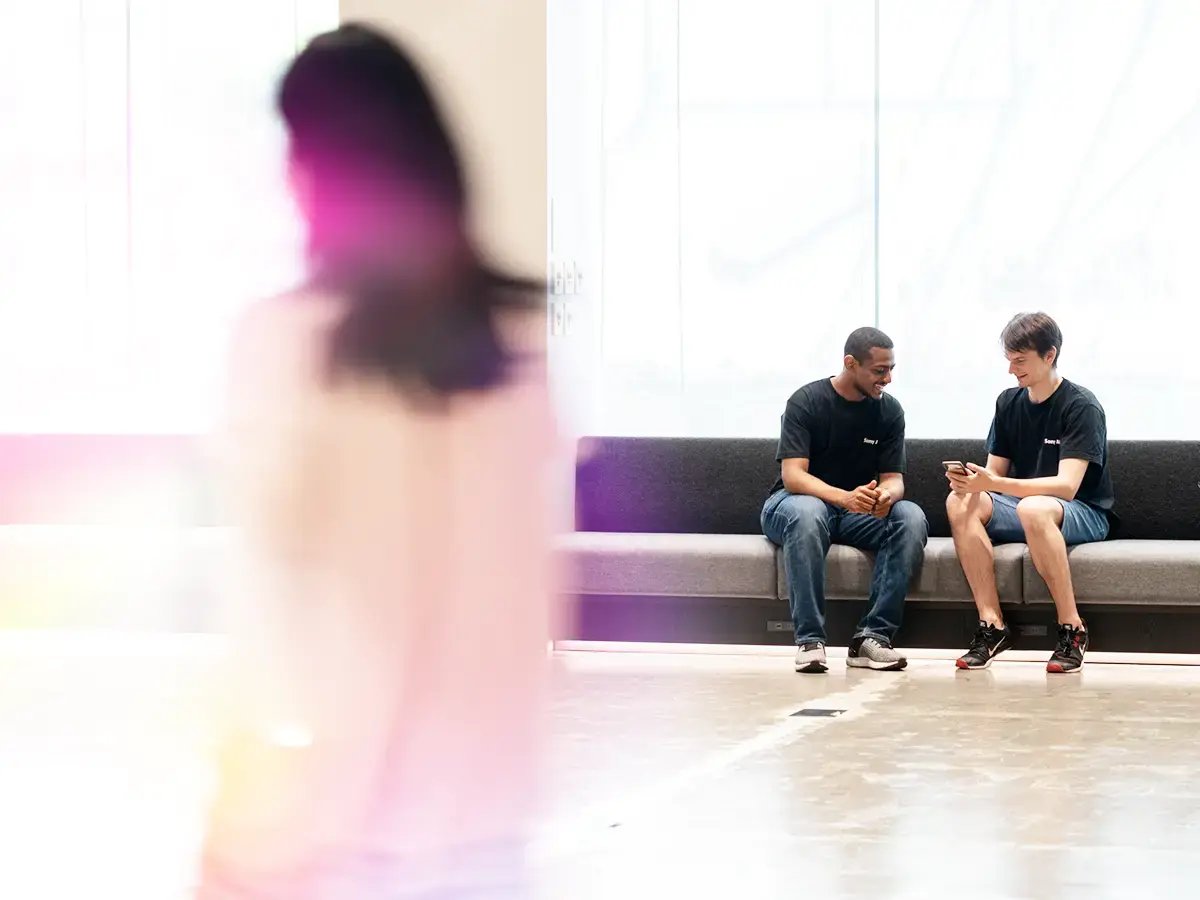 A blurred figure walks past two people sitting on a bench, who are engaged and smiling while looking at a smartphone