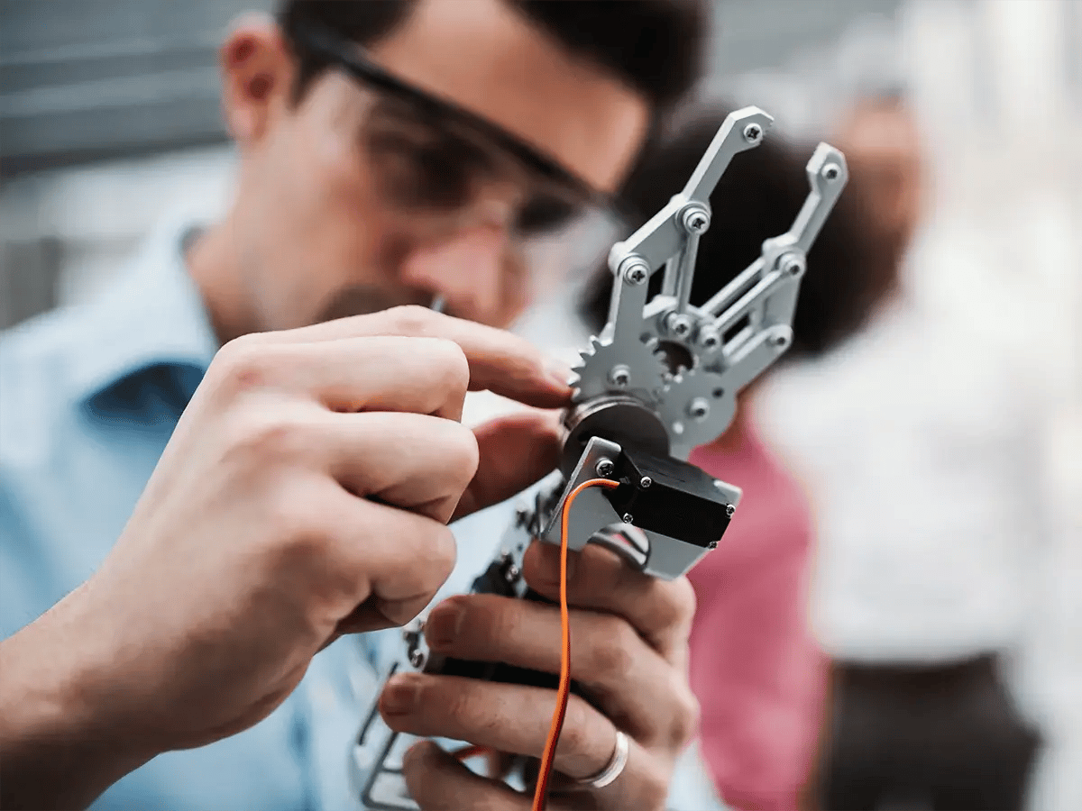 A focused individual works on a robotic hand with intricate metal parts and orange wires