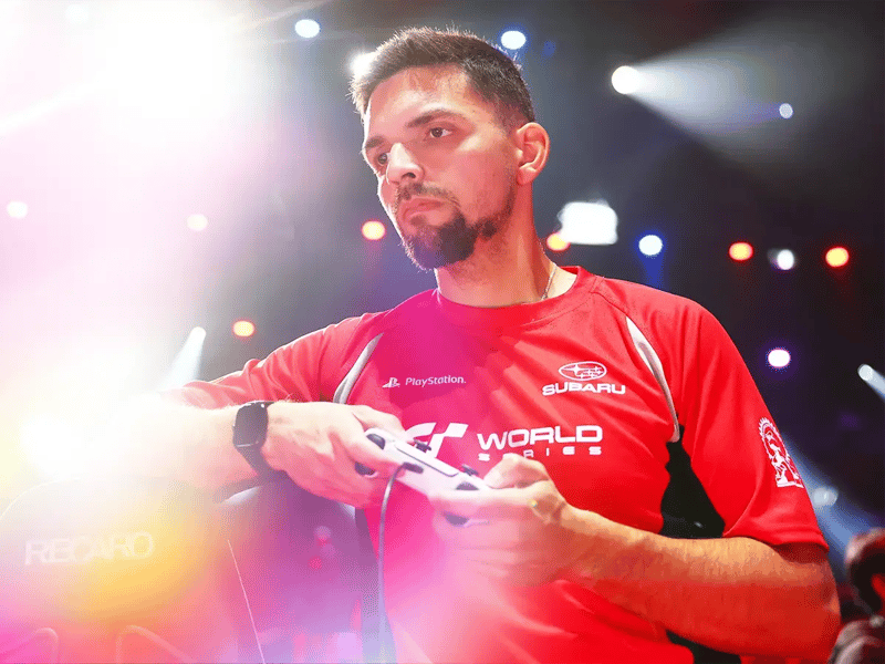 A focused person in a red Subaru and PlayStation shirt holds a game controller under colorful stage lights
