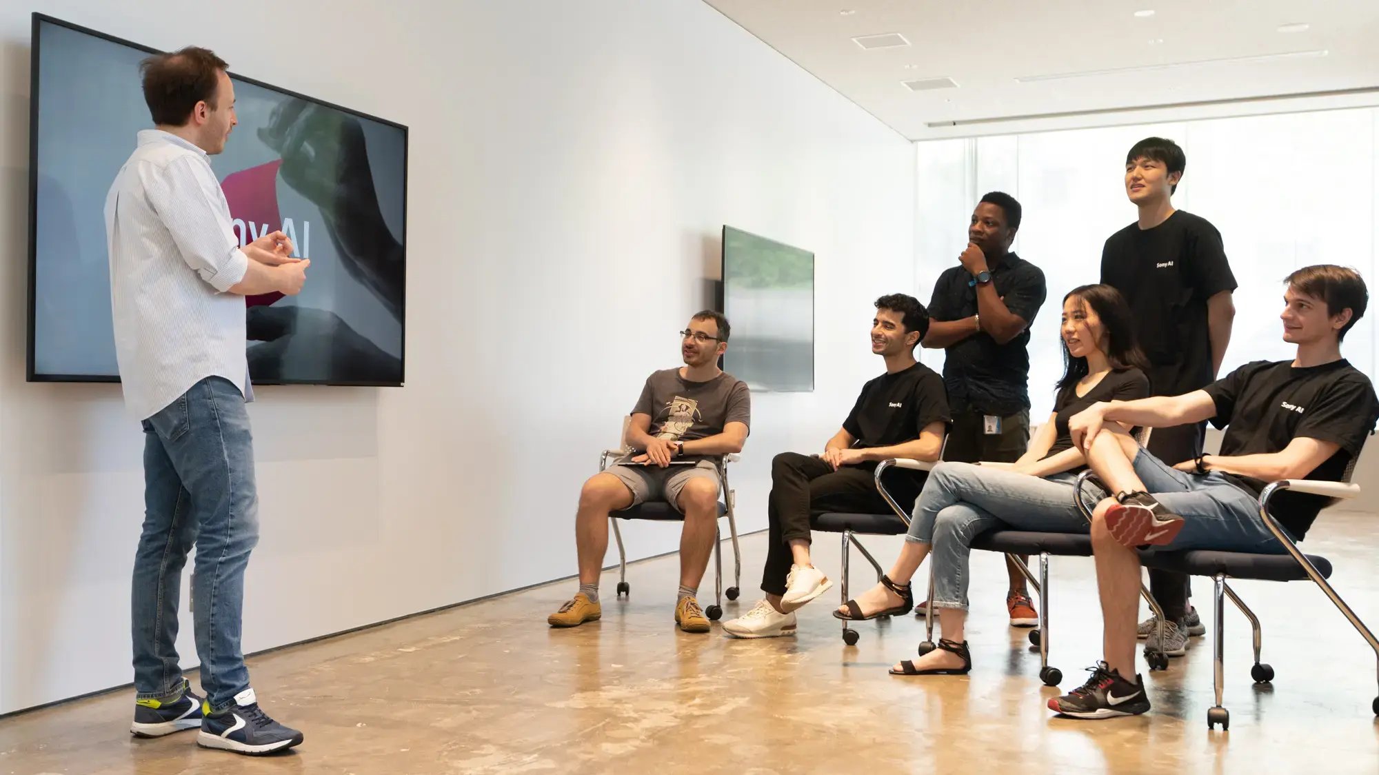 A group of six people in casual attire attentively watches a person speaking near a monitor in a modern, bright room