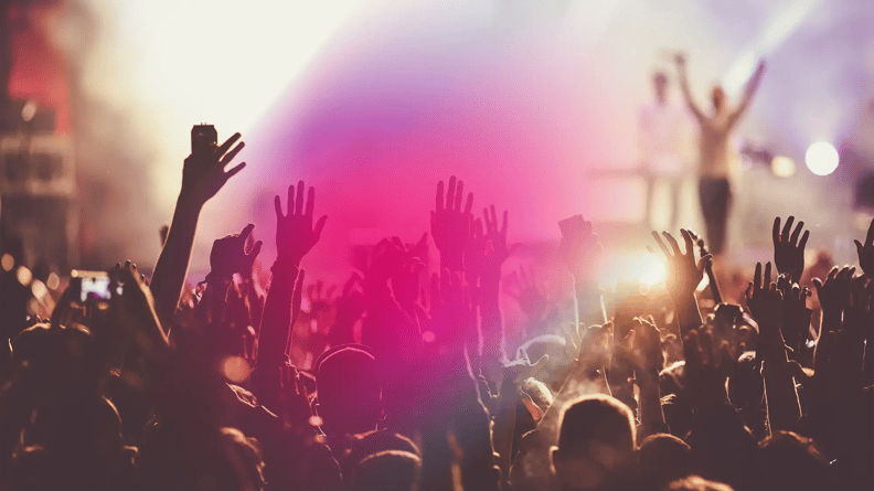 A lively concert scene with a crowd of raised hands silhouetted against bright stage lights, creating an energetic and euphoric atmosphere