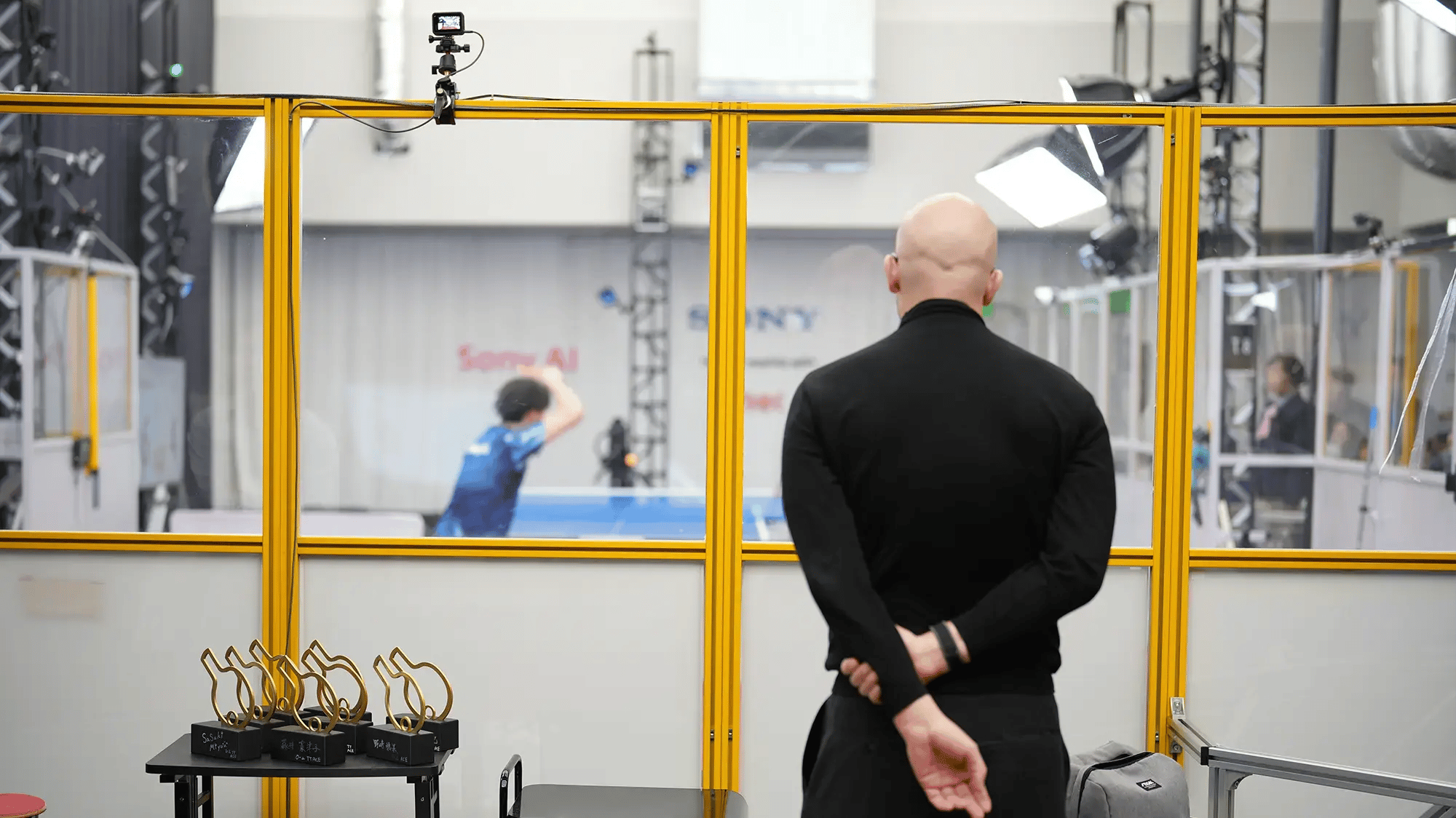A man in black watches a ping pong match through a glass panel