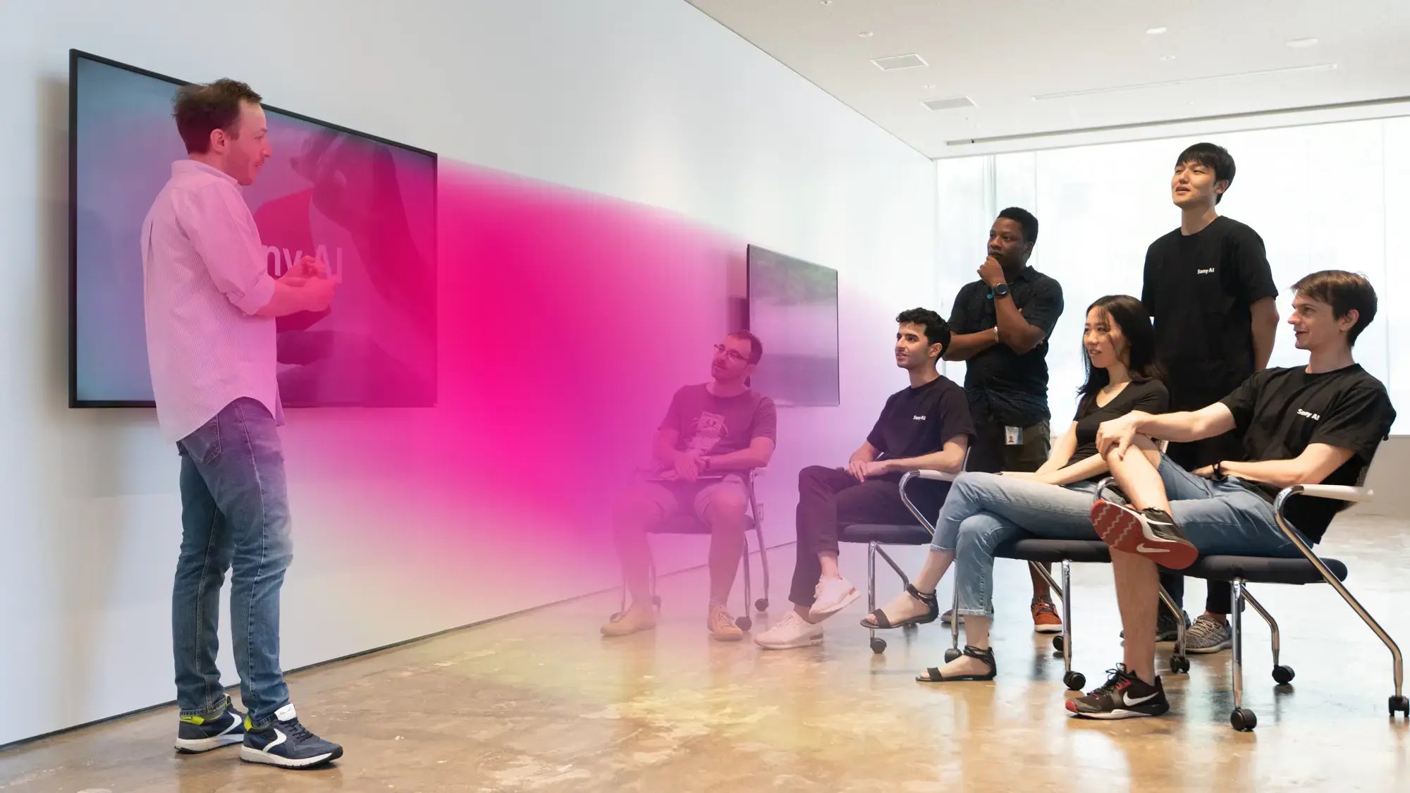 A man presents to six seated, engaged individuals in a bright room with floor-length windows