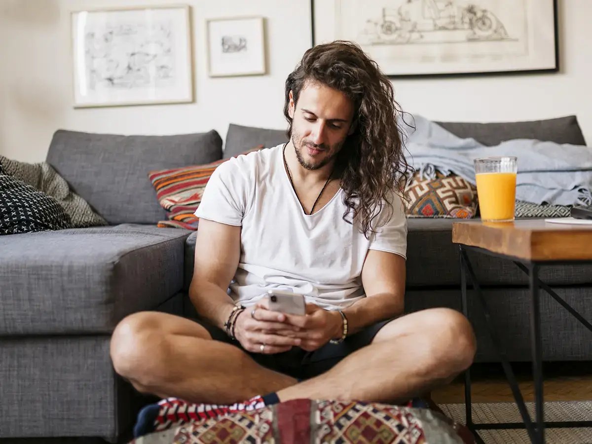 A man with long hair sitting cross-legged on the floor, using a smartphone