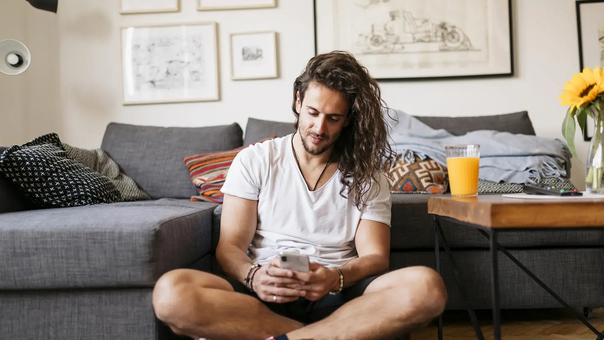 A man with long hair, wearing a white T-shirt, sits on the floor using his phone in a cozy living room