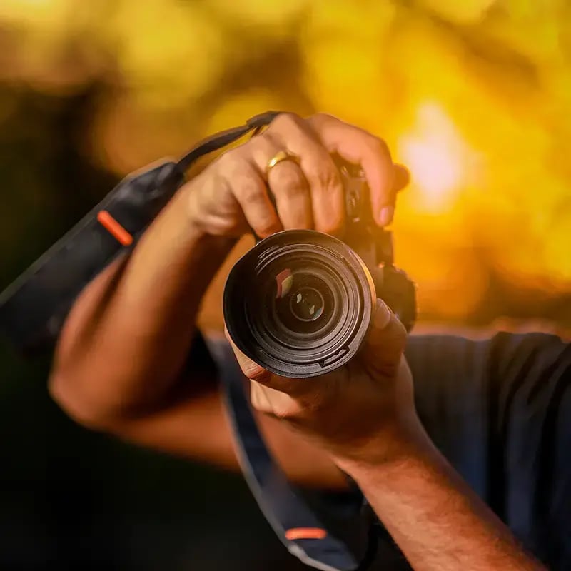 A person holds a camera close to the lens, focused against a warm, blurred sunset background