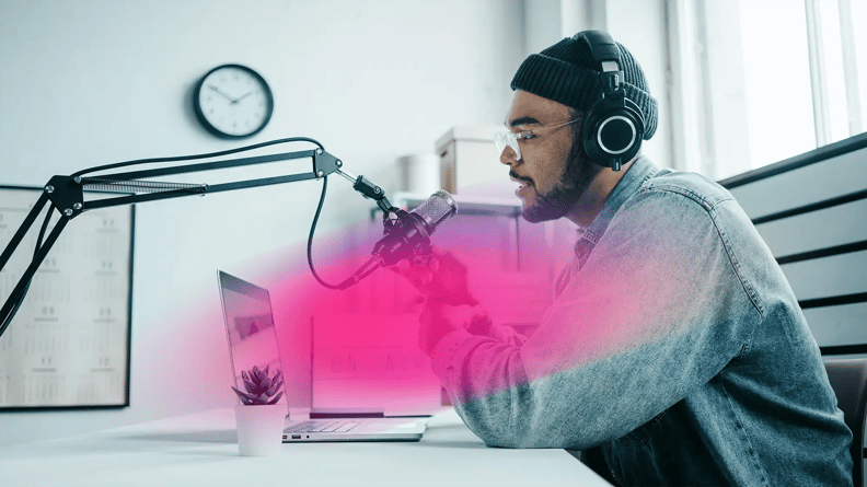 A person in headphones speaks into a microphone, seated at a desk with a laptop
