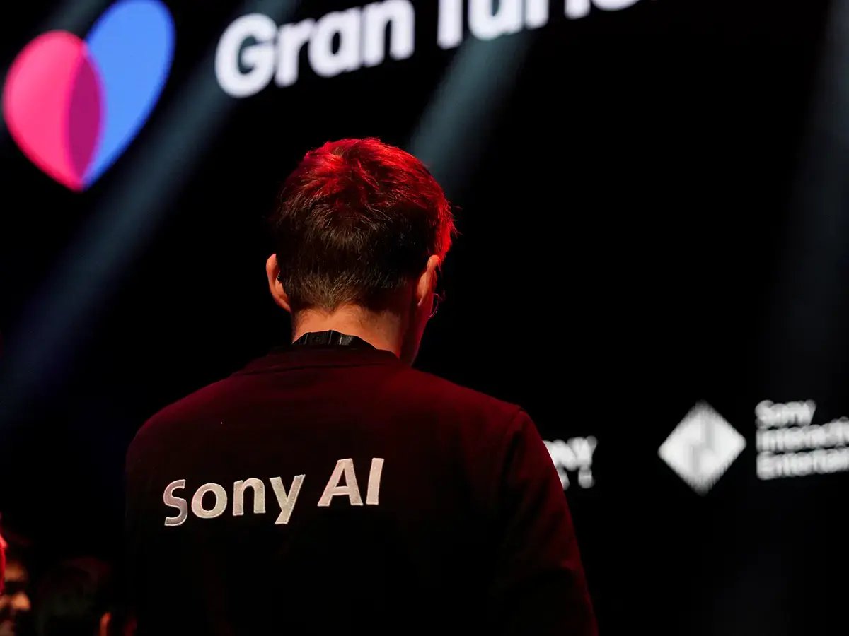 A person wearing a Sony AI shirt faces a screen displaying the words Gran Turismo with a heart graphic