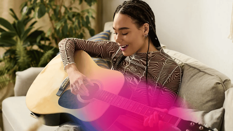 A person with braided hair sits on a couch, playing an acoustic guitar with a joyful expression