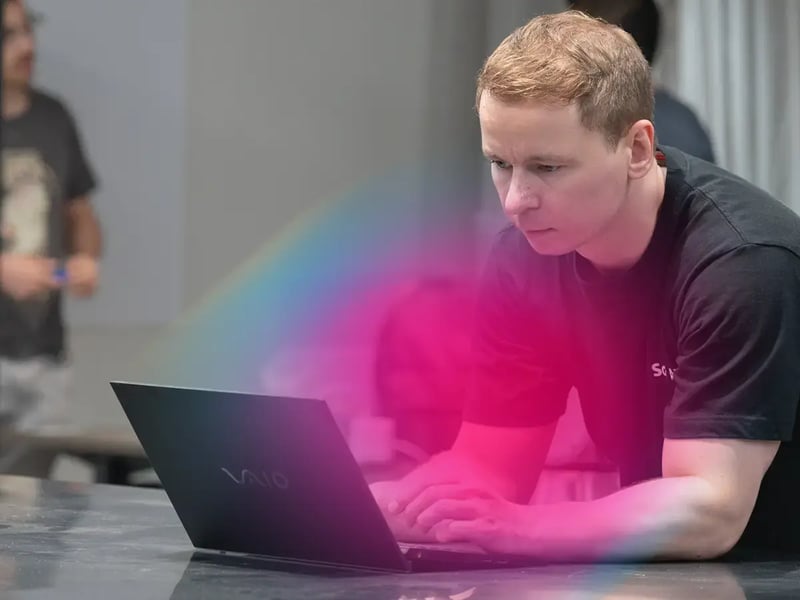 A person with short hair focuses intently on a laptop, surrounded by a pink hue