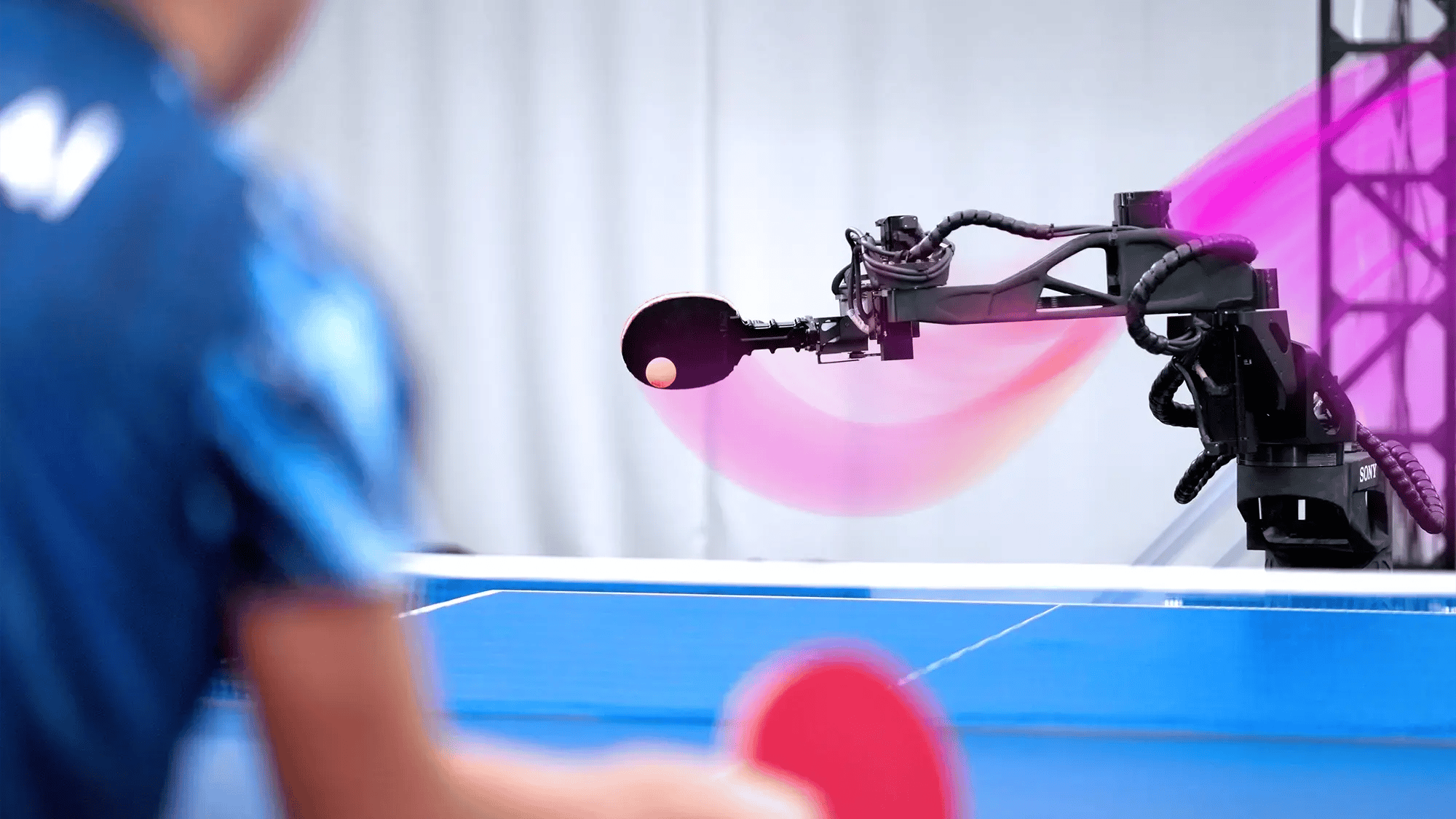 A robot arm, set against a white backdrop, is playing table tennis with a person in a blue shirt