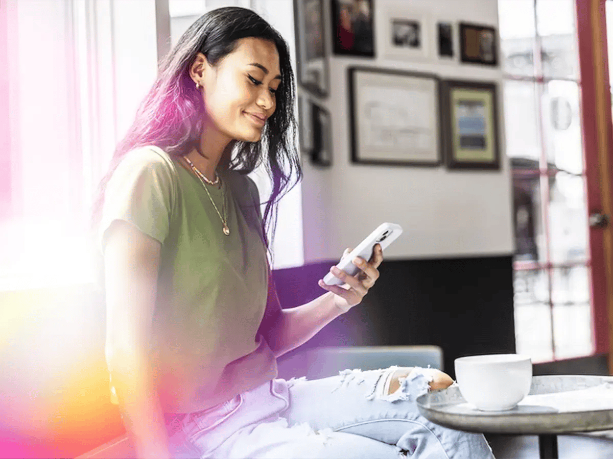 A woman in a green shirt sits in a bright café, smiling at her phone