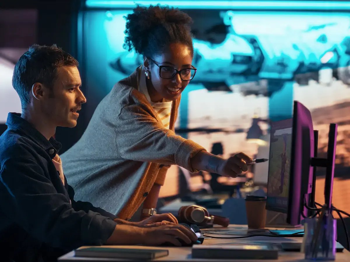 A woman points at a computer screen while a man looks on, both in an office with a blue-lit background