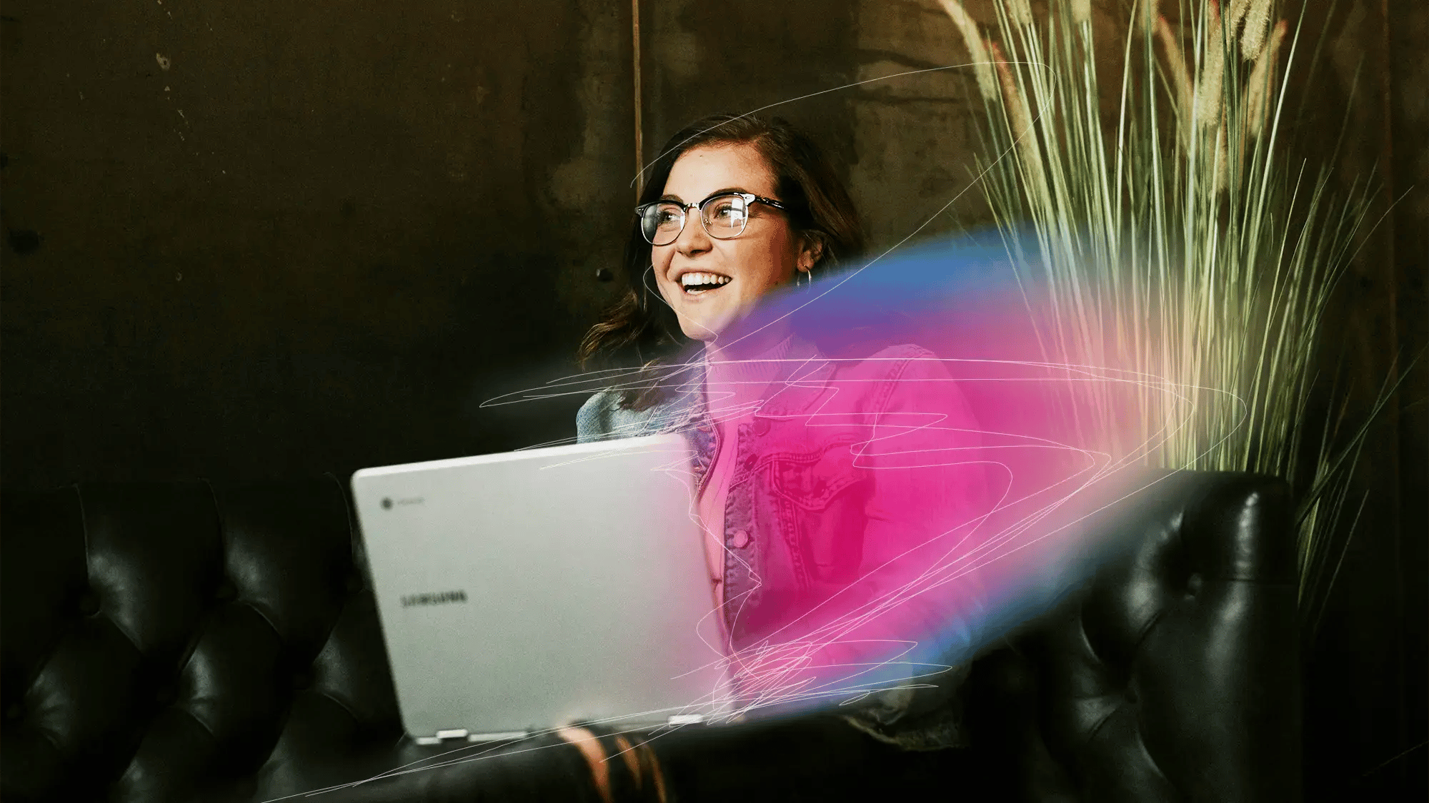 A woman with glasses and a pink jacket smiles while using a laptop on a dark sofa