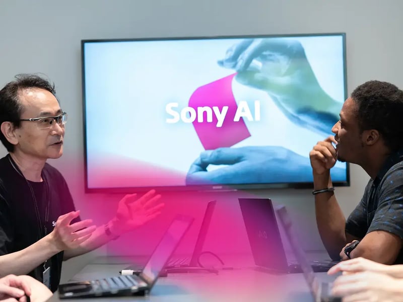 Two people sit at a table discussing in front of a screen displaying Sony AI with a vibrant pink graphic
