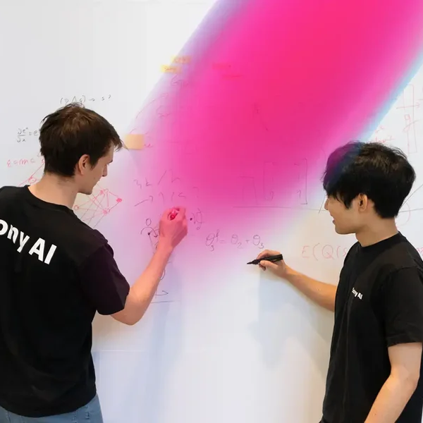 Two people in black Sony AI shirts collaborate at a whiteboard, discussing equations and diagrams, with a bright pink gradient overlay