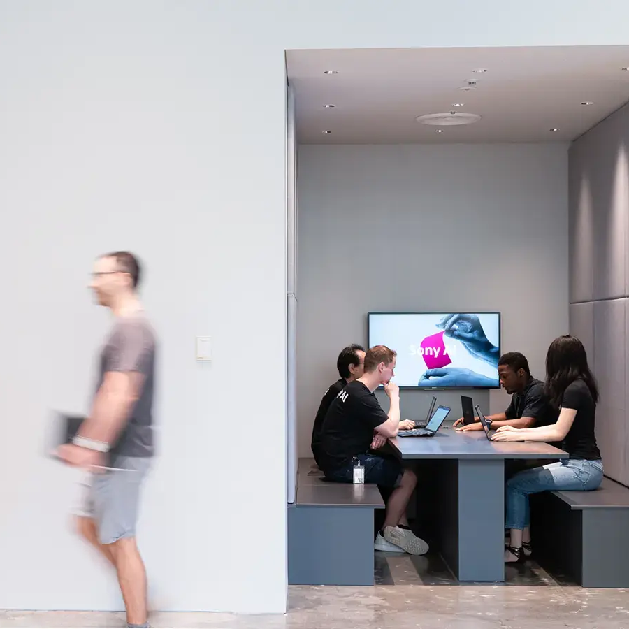 A group of four people sits at a modern conference table in a small, enclosed space, collaborating on laptops