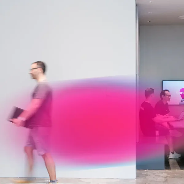 A blurred man walks past a meeting room, its vibrant pink motion visible. Inside, four people sit at a table with laptops, focused on a screen displaying Sony.